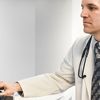Medical professional wearing white coat and stethoscope reviews medical images on a desktop computer.