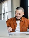 A person with a pen in hand smiles and looks at a laptop.