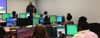 Adult stands in front of multiple young people at computers in classroom setting.