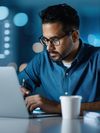 A focused man with a beard and glasses works on a laptop in an office, with a blurred graph in the background.