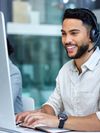 A smiling man wearing a headset, typing on the keyboard of a computer workstation in an office environment.