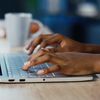 Hands work on a laptop with a notebook and coffee mug in the background.