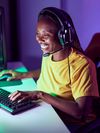 An African-American woman is at her desk playing video games while wearing a gaming headset.