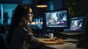 A bespectacled, brunette woman is sitting at her workspace and looking at her laptop in a dimly lit office.