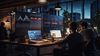 Two workers in a dimly lit office are looking at their laptops in front of two big screens showing data visualizations or dashboards.