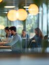 Several young professionals sit at separate tables in an open co-working space, working individually on laptops.