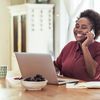 Person in red button-down talks on a cell phone and works on a laptop at a home desk.