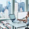 Person sits at desk working on computer in front of large window overlooking urban city.