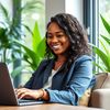 A smiling woman typing on her laptop keyboard sitting in a well-lit office with green potted plans in teh background.