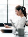 Person in casual business attire sits in an office setting and looks at a handheld device. 