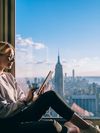A blonde woman wearing glasses reads a tablet by a window overlooking the New York City skyline, with the Empire State Building visible in the background.