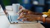 Hands using a laptop computer with a notebook and coffee mug in the background.