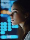 A young woman sits in a dimly-lit room, focusing on several digital  screens displaying colorful graphs and charts. The screen's glow reflects on her face as she analyzes the data, emphasizing a data-driven environment.
