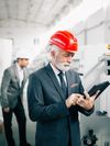 Person wearing a suit and a hardhat looks at a tablet while inspecting equipment.
