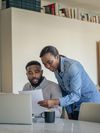 Two people look over paperwork. One is standing and leaning over the seated person?s shoulder.