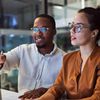Two coworkers sit at a desktop computer and explore the differences in AI and machine learning.