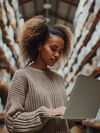 A young woman stands in a warehouse filled with stacked large cardboard boxes on shelves. She is holding and working on a laptop, appearing focused as she likely manages inventory or tracking shipments.