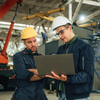 Two construction workers in hard hats and jackets reviewing a laptop inside an industrial warehouse.